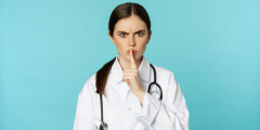 A healthcare worker in a white coat makes a "shhhh" finger over their mouth and looks at the camera with concern.