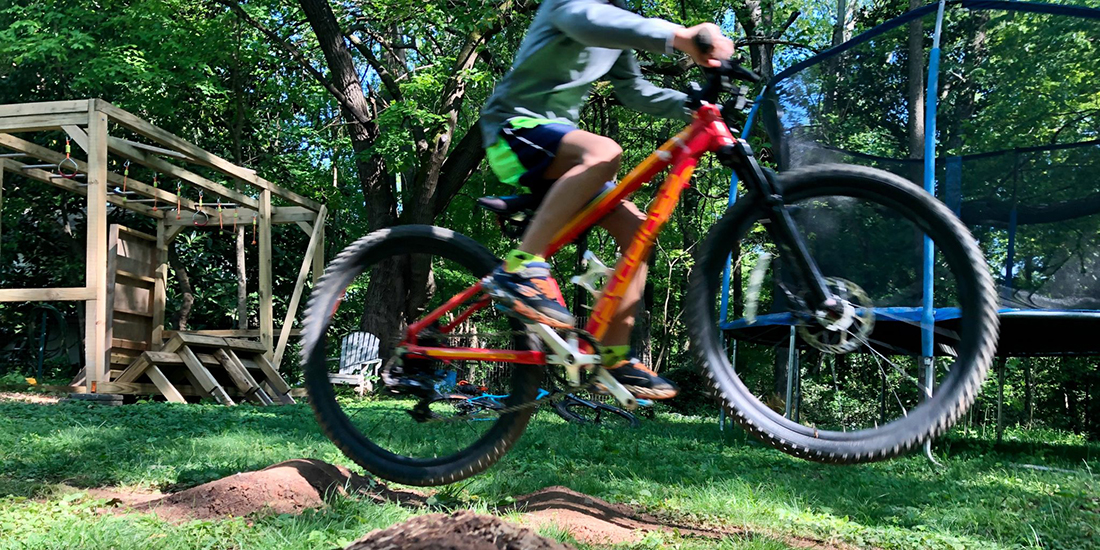 A kid jumps a mountain bike over a ramp in a sunny backyard.