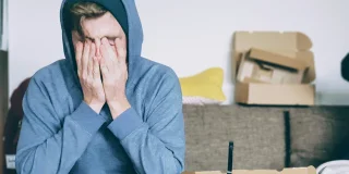 A young guy in a blue hoody sits on a couch with his head in his hands. There are empty boxes around him.
