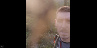 A hiker talks into his phone camera while a mosquito accosts the screen.