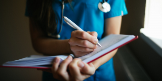 A doctor in a bright blue uniform scribbles a note into a serious looking notebook.