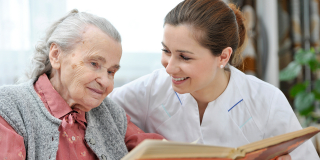 A younger homecare worker reads a newspaper with an elderly woman.