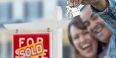 Two people smile in front of a sold sign. One is holding a set of house keys.