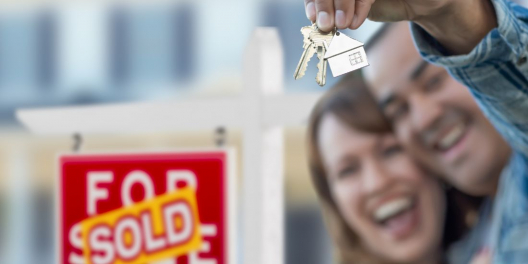 Two people smile in front of a sold sign. One is holding a set of house keys.